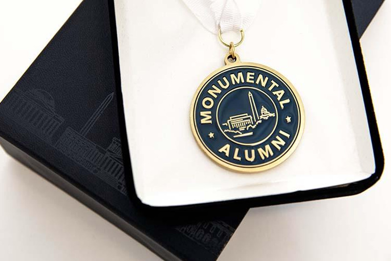 A blue and gold amulet with the words "Monumental Alumni" surrounding an icon of Washington, DC monuments sits in a small case on a white background